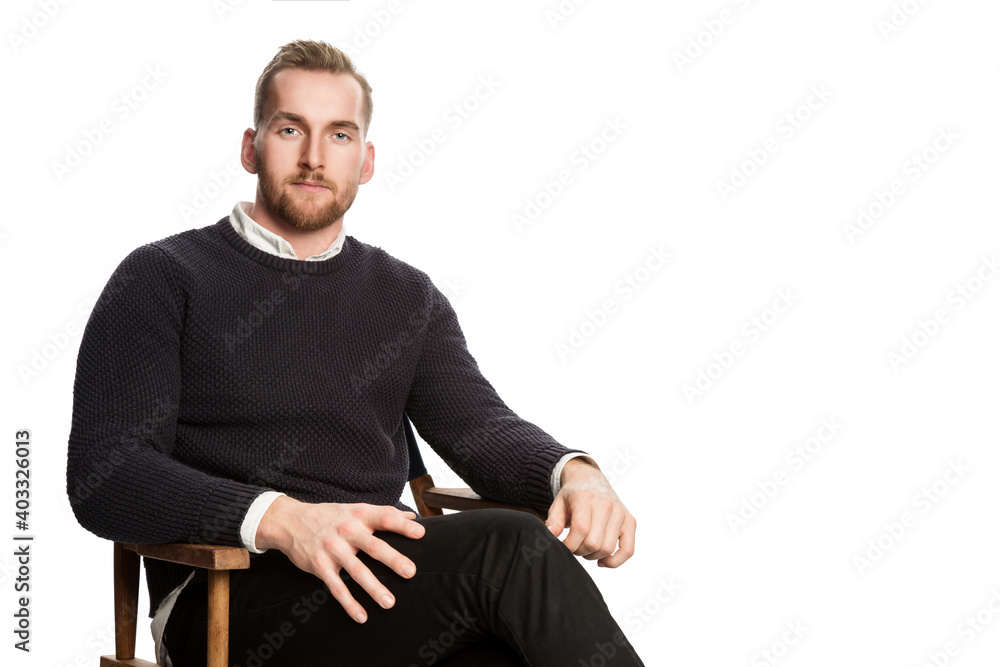 Attractive blonde man sitting down in front of a white background. Wearing dark pants and a dark sweater. Attractive blonde man sitting down in front of a white background. Wearing dark pants and a dark sweater.