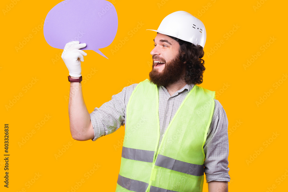 A cheerful young bearded architect is looking at the speech bubble that he is holding near a yellow wall wearing a helmet some gloves and a phosphorescent vest .