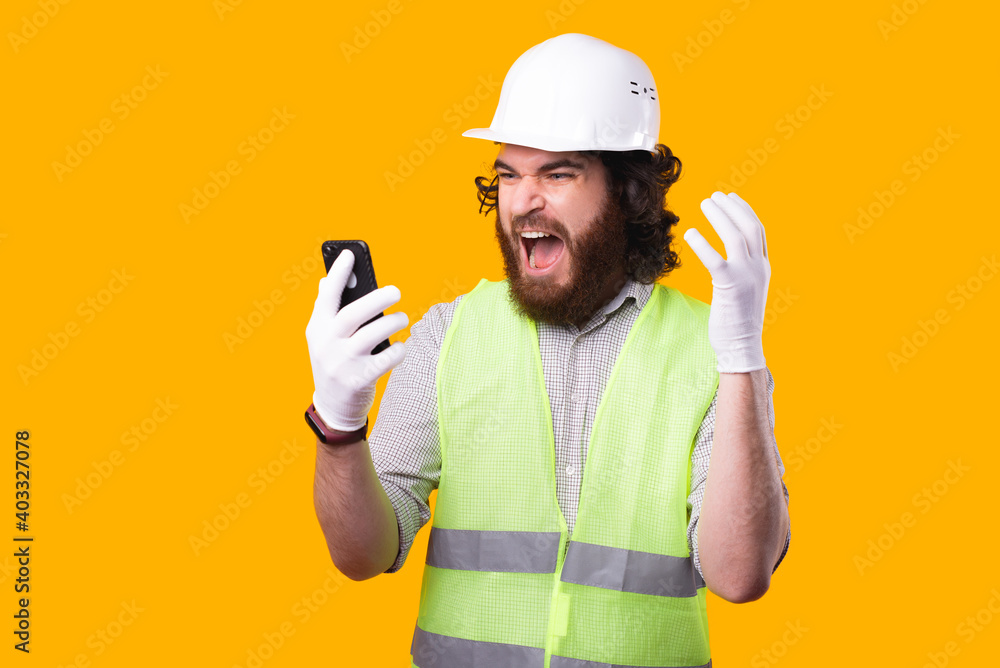 A bearded young engineer is looking shocked at his phone near a yellow wall . A bearded young engineer is looking shocked at his phone near a yellow wall .