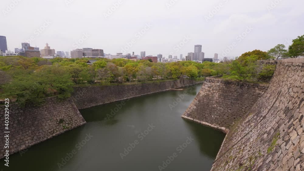 Panoramic view of old moat around Osaka Castle, green trees around, impressive large stone walls and water dyke. Modern city buildings seen on background Panoramic view of old moat around Osaka Castle, green trees around, impressive large stone walls and water dyke. Modern city buildings seen on background