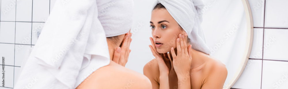 displeased young woman with towel on head looking in mirror and touching face in bathroom, banner displeased young woman with towel on head looking in mirror and touching face in bathroom, banner