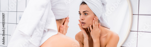 displeased young woman with towel on head looking in mirror and touching face in bathroom, banner
