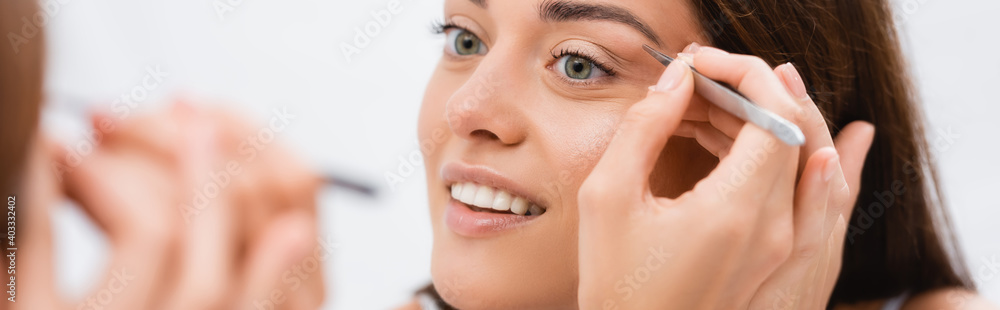 smiling woman tweezing eyebrows near mirror, blurred foreground, banner