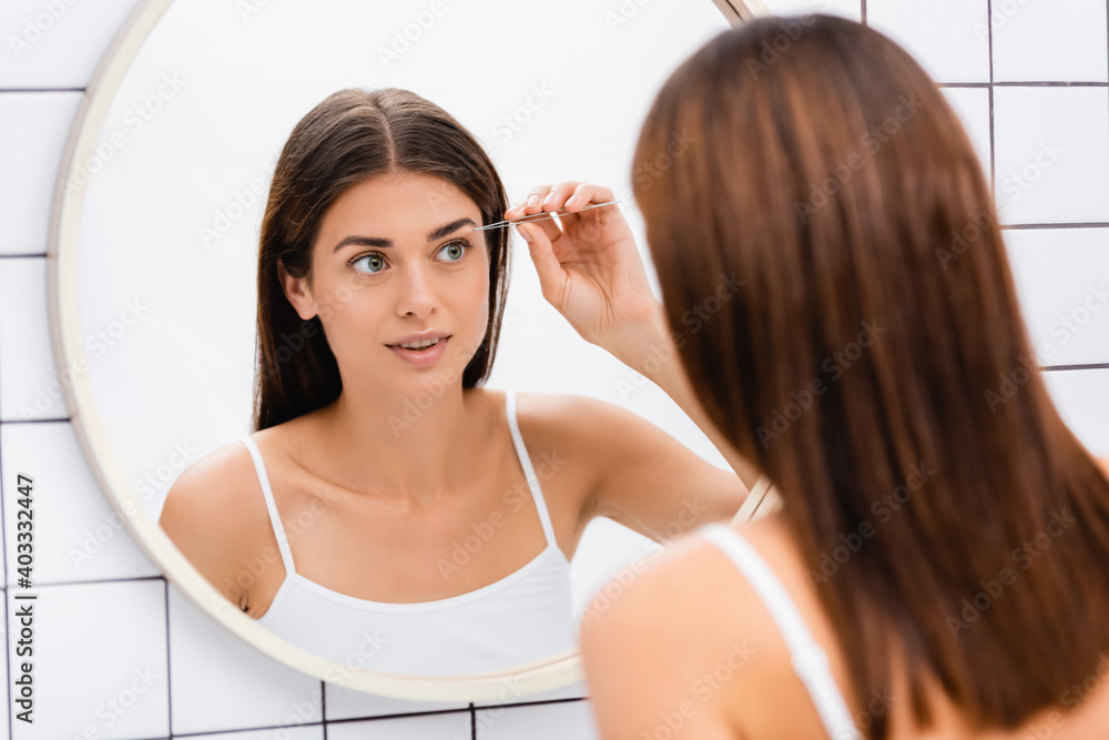 young woman in white singlet tweezing eyebrows near mirror in bathroom young woman in white singlet tweezing eyebrows near mirror in bathroom