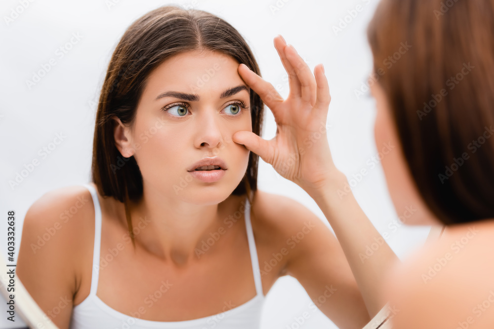 worried young woman looking in mirror and touching face, blurred foreground worried young woman looking in mirror and touching face, blurred foreground