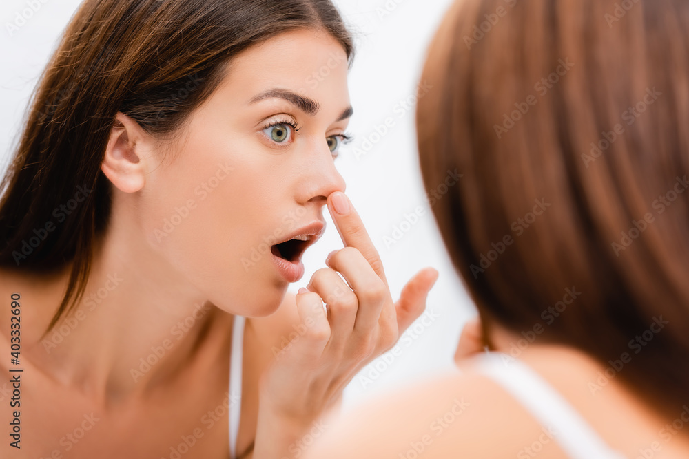 shocked young woman touching nose while looking in mirror in bathroom, blurred foreground shocked young woman touching nose while looking in mirror in bathroom, blurred foreground