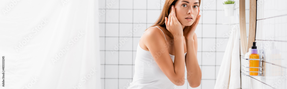 displeased young woman touching face while looking at camera in bathroom, banner displeased young woman touching face while looking at camera in bathroom, banner