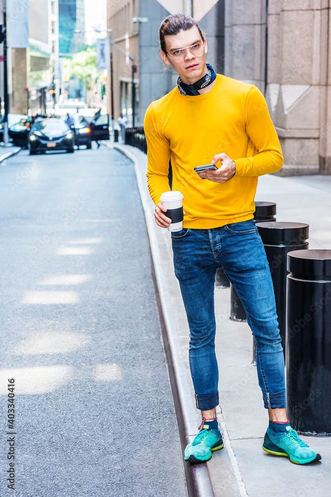 Φωτογραφία Young Hispanic American with hair bun, wearing glasses, yellow long sleeve T shirt, blue jeans, green sneakers, holding cup of coffee, standing on street in New York City, texting on cell phone