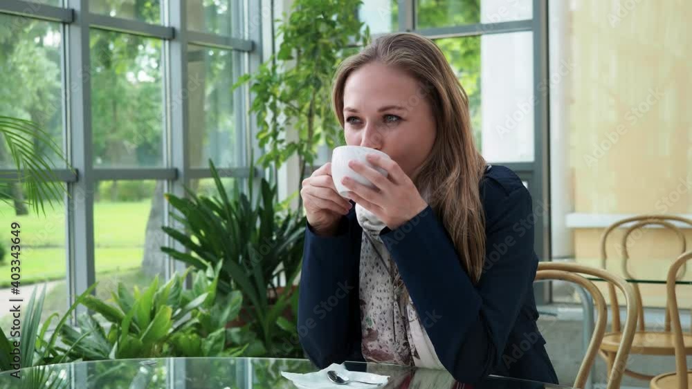 Gorgeous young long hair blonde woman wearing blue cloak enjoying a cup of coffee sitting at glass table near the window in cozy cafe day time Gorgeous young long hair blonde woman wearing blue cloak enjoying a cup of coffee sitting at glass table near the window in cozy cafe day time