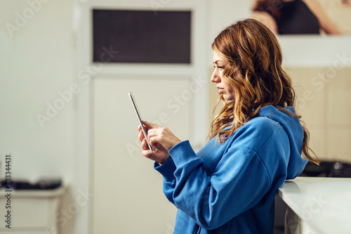 Side view portrait of beautiful caucasian woman leaning on the desk at work looking to smartphone - Brunette female using mobile phone at online app at home for sms or selfie