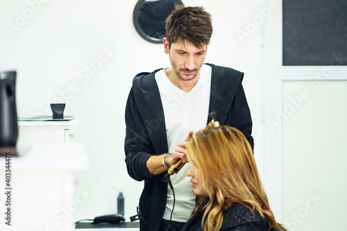 Young man hairdresser at work with adult woman using hair iron to straighten the hair of customer client - copy space
