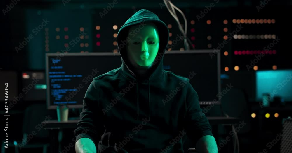 Wanted Hacker with Masked Face looks into the Camera. In the Background His Operating Room with Multiple Displays and Cables. Anonymous man covers her mouth with her finger in a gesture of silence Wanted Hacker with Masked Face looks into the Camera. In the Background His Operating Room with Multiple Displays and Cables. Anonymous man covers her mouth with her finger in a gesture of silence