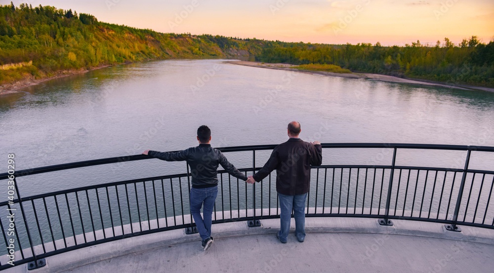 male gay couple holding hands, on a bridge looking at the horizon, over a river of blue water and pine trees in the background, on an autumn afternoon