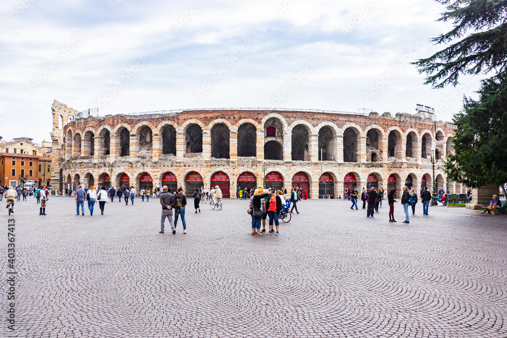 Tourists walking around the Piazza Bra square on a cloudy day, inspect the Arena and take pictures against its background in Verona, Italy.