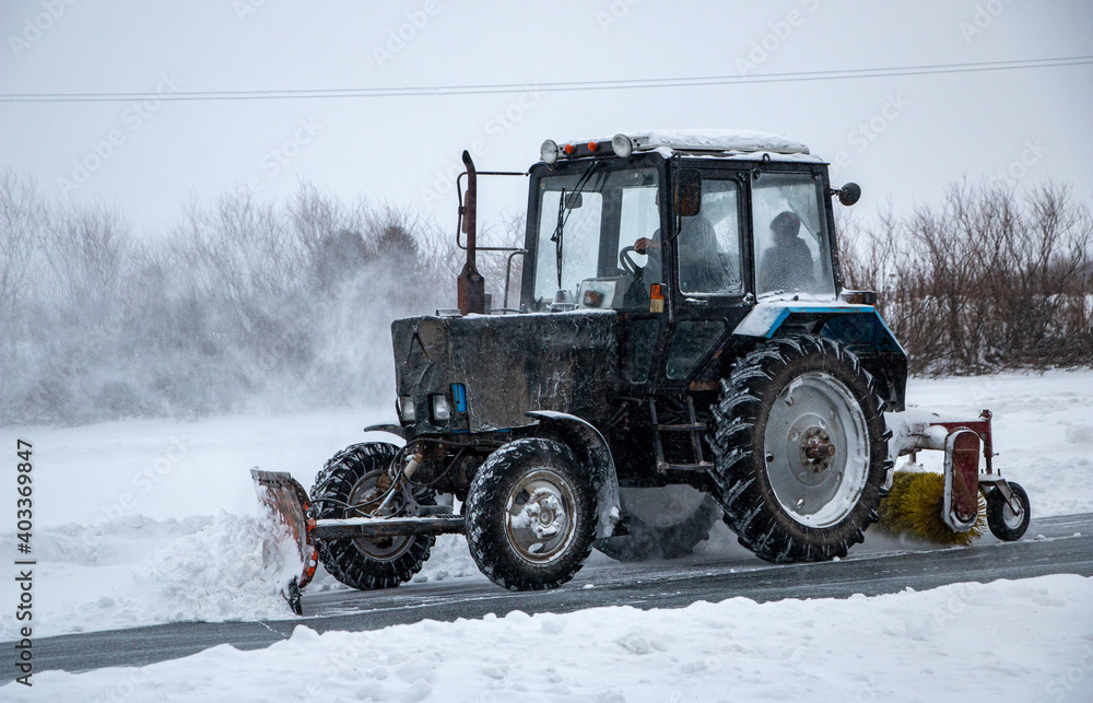 an old snow-covered tractor with blade removes snow from the road, sweeps snow from the road with large brush, in severe blizzard. an old snow-covered tractor with blade removes snow from the road, sweeps snow from the road with large brush, in severe blizzard.