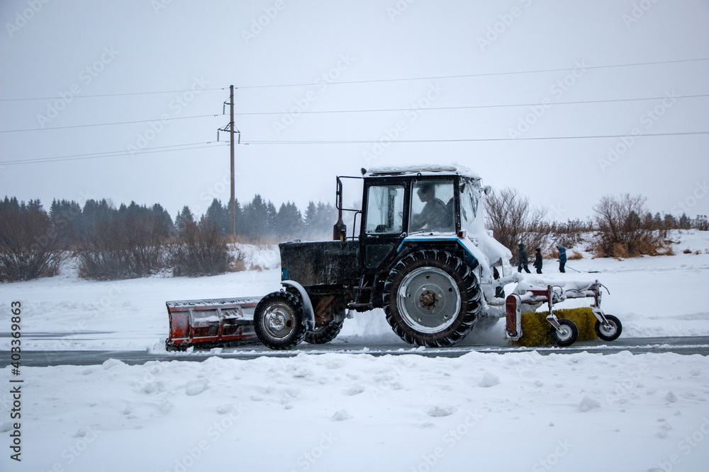 an old snow-covered tractor with  blade removes snow from the road, sweeps snow from the road with  large brush, in  severe blizzard.