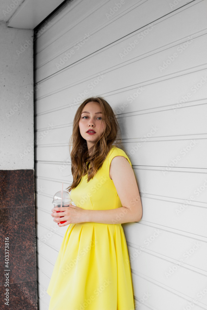 Fototapeta premium Young woman in illuminating yellow dress trend color of 2021 standing in front of white wall, holding glass of lemonade and looking at camera. Girl having fun. Urban style and fashion. Casual outfit