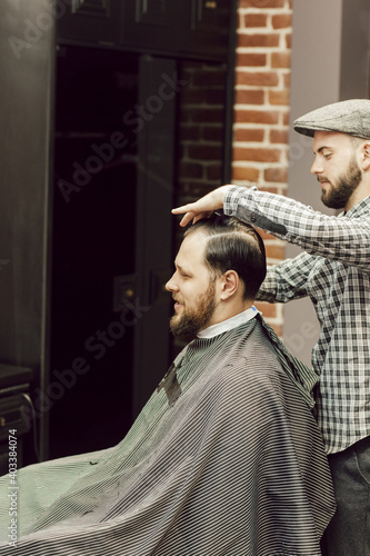 Young bearded man getting haircut while sitting in chair at barbershop. The hairdresser cuts and combs client's hair with a comb. Young men in a barbershop