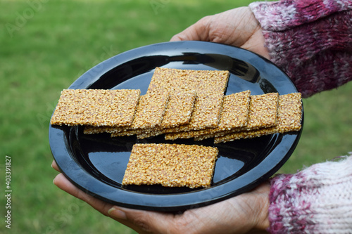 Female hand holding Winter special sweet dish of White sesame and jaggery melted in clarified butter and rolled in to sheets garnished and decorated with fresh leaves on black plate photo