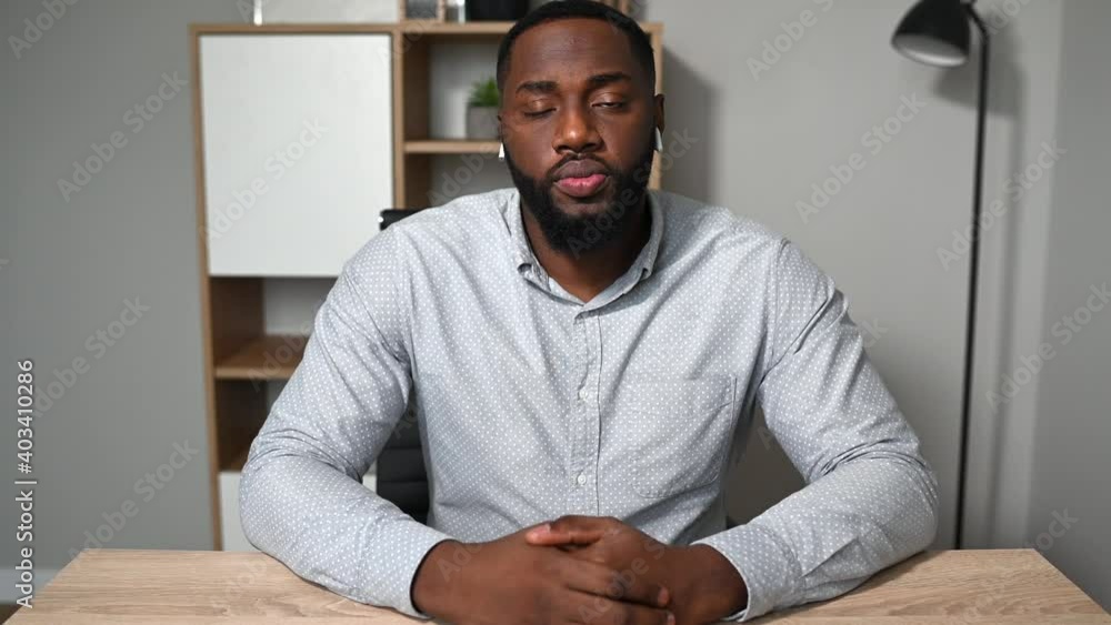 A young African man in smart casual shirt looks at webcam with a serious face and then smiles, online meeting in office. Video call, online interview concept A young African man in smart casual shirt looks at webcam with a serious face and then smiles, online meeting in office. Video call, online interview concept