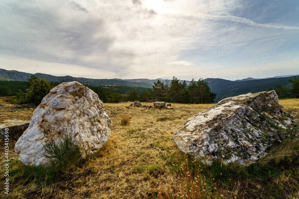 Landscape of rocks with green plants, dramatic sky with clouds and distant mountains.
