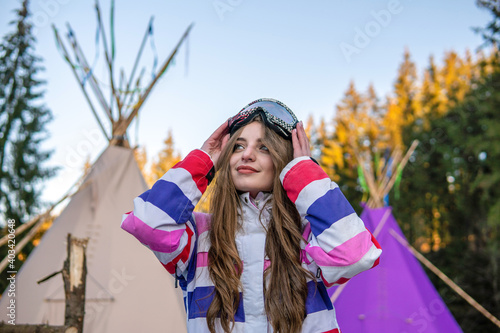 Portrait of Pretty Caucasian young woman Near The Indian Tipi or Wigwam in ski outfit and Winter Sports Mask on her Head. Portrait of cheerful blond woman at ski resort
