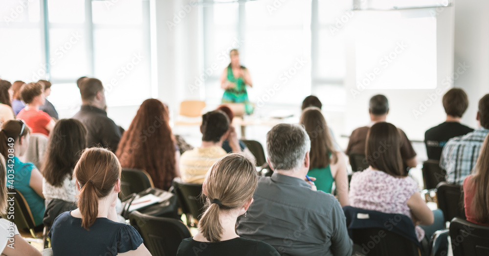 Fototapeta premium Business and entrepreneurship symposium. Female speaker giving a talk at business meeting. Audience in conference hall. Rear view of unrecognized participant in audience.