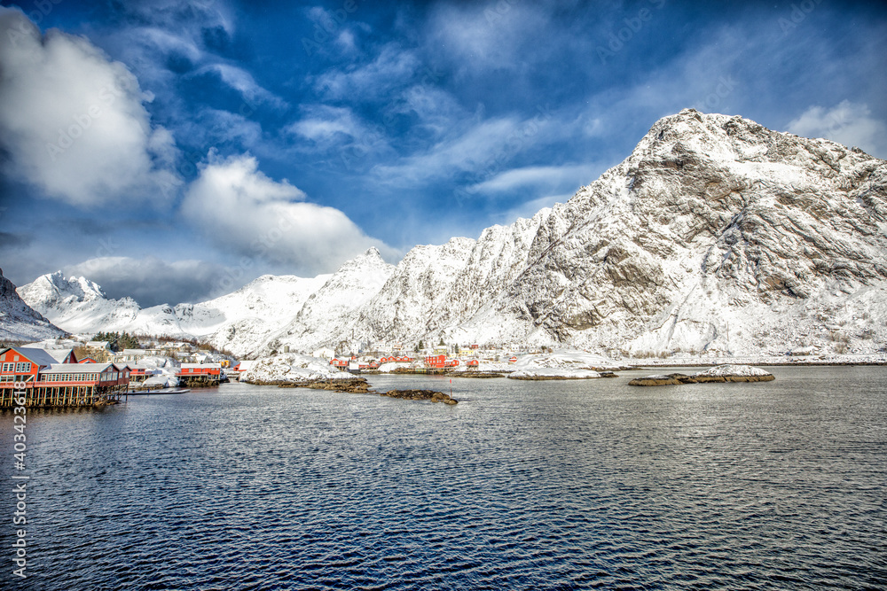 Norway. Lofoten. The village is on the water in the background of white mountains and blue sky.