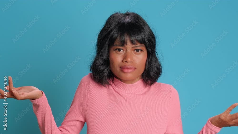 Attractive hesitant dark skinned Asian American woman spreads hands sideways as being clueless and doubtful cannot make choice poses unaware dressed in casual pink jumper isolated over blue background Attractive hesitant dark skinned Asian American woman spreads hands sideways as being clueless and doubtful cannot make choice poses unaware dressed in casual pink jumper isolated over blue background