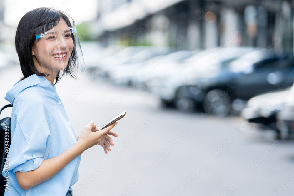 Young Asian woman wearing face shield and holding a mobile phone standing at a domestic street. Concept commuters in covid time Young Asian woman wearing face shield and holding a mobile phone standing at a domestic street. Concept commuters in covid time