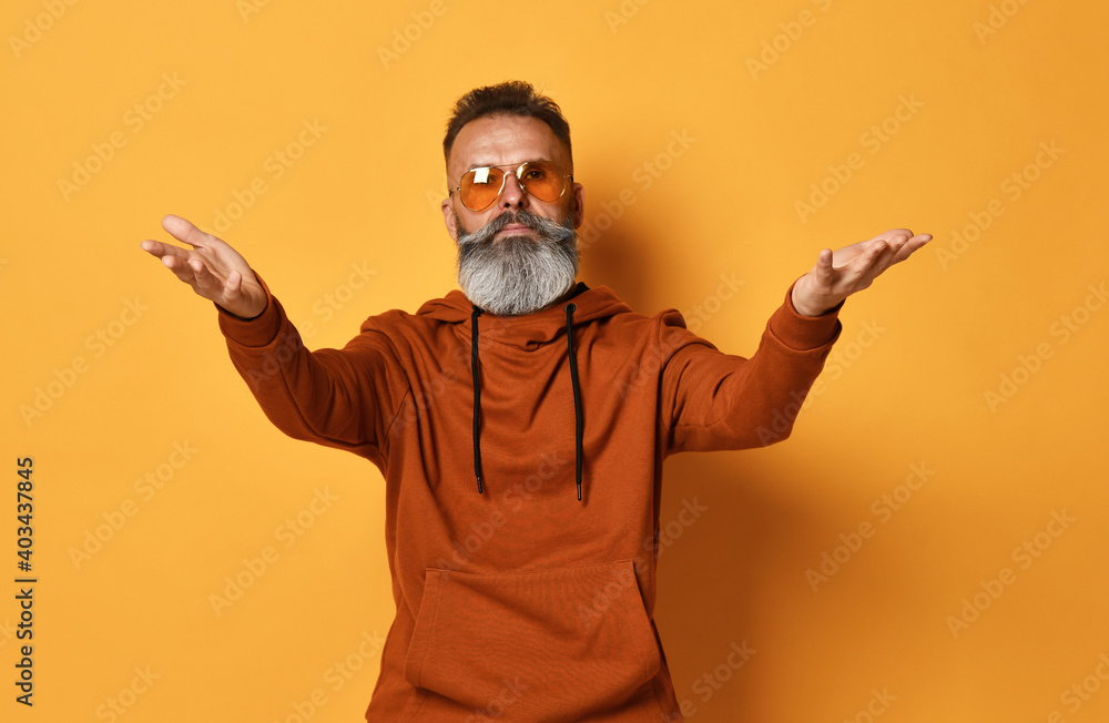 Serious brutal handsome grey-haired bearded man wearing casual sportswear, trendy sunglasses stretching hands to camera. Welcome, wide open hug. Indoor studio shot isolated on yellow background