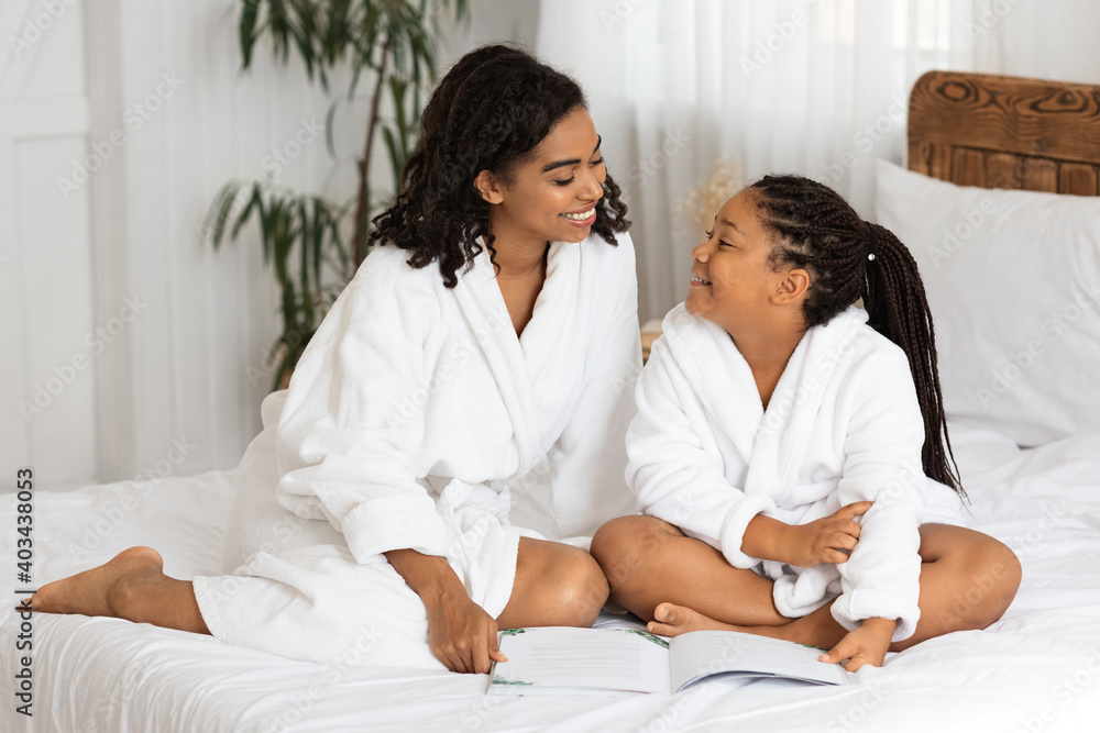 Cheerful African Girl And Her Mother In Bathrobes Reading Book On Bed Cheerful African Girl And Her Mother In Bathrobes Reading Book On Bed
