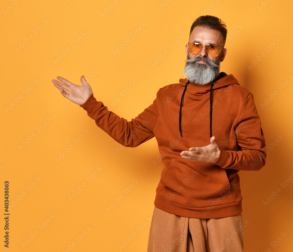 Serious brutal handsome grey-haired bearded man wearing casual sportswear pointing to left looking at camera standing isolated on yellow background with copy space. Studio shot. Sportive fashion style