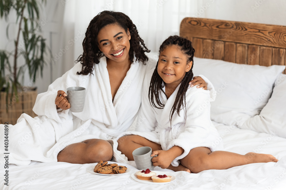 Joyful Black Mom And Daughter In Bathrobes Eating Snacks In Bed