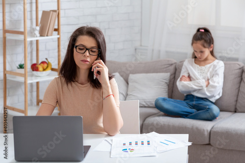 Young mother working online, using laptop and phone, upset child sitting on sofa nearby, suffering from neglect