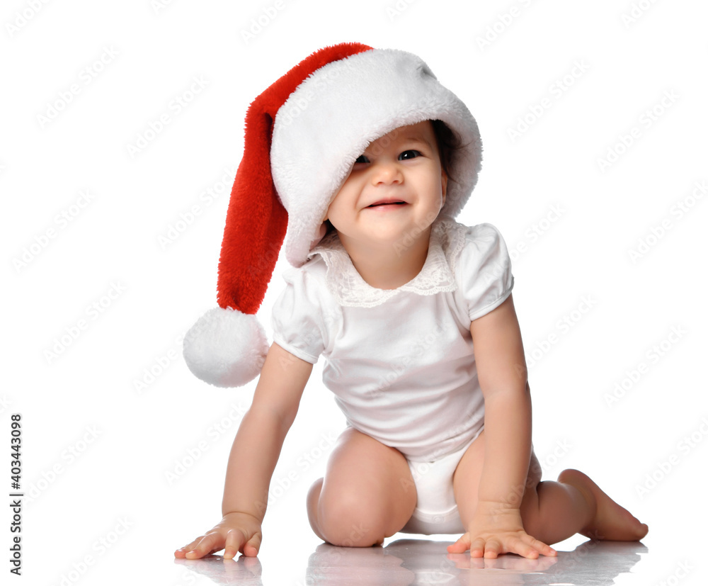 Cute funny smiling little baby girl in overalls and red Santa Claus hat sitting on a white floor studio background. Isolated portrait shot. Merry christmas, happy new year concept Cute funny smiling little baby girl in overalls and red Santa Claus hat sitting on a white floor studio background. Isolated portrait shot. Merry christmas, happy new year concept