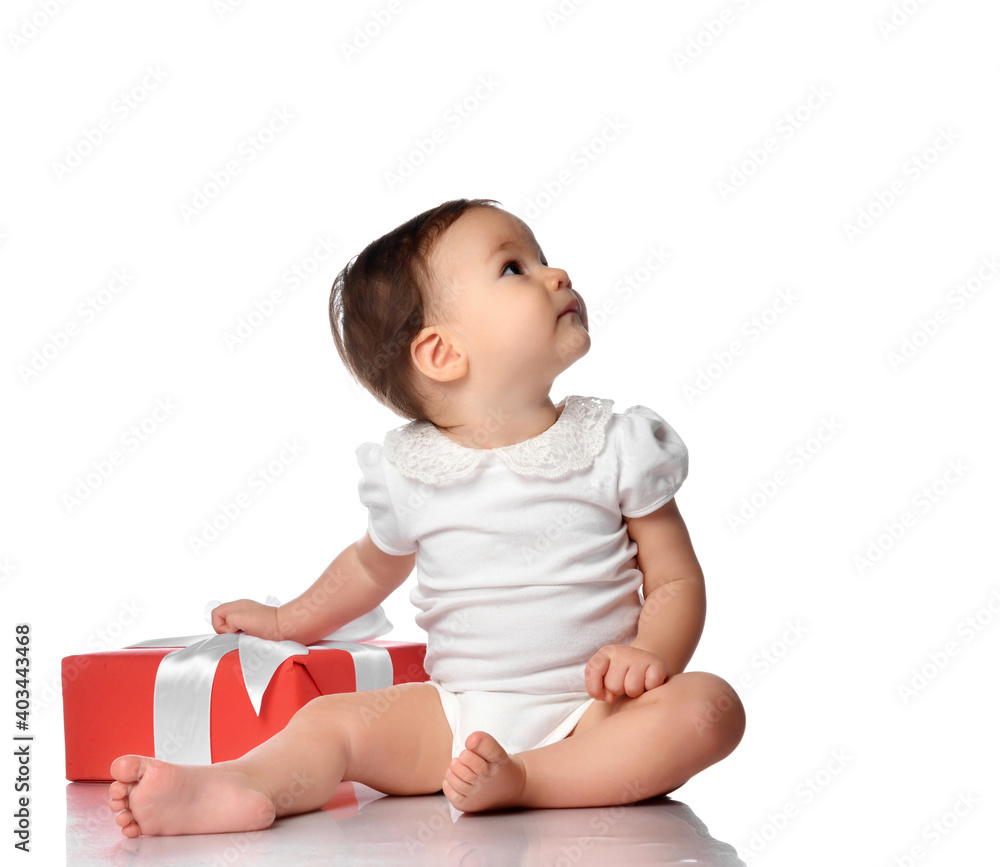 Baby and wrapped festive gift box with bow. Surprised infant girl child wearing jumpsuit sitting on white floor studio background looking aside. Isolated portrait. Birthday or Christmas time concept
