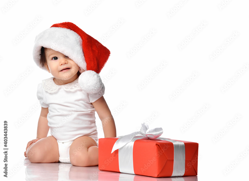 Cute little baby girl in jumpsuit and red Santa Claus hat, sitting on white floor studio background with wrapped holiday gift box. Isolated portrait shot. Christmas, New Year concept Cute little baby girl in jumpsuit and red Santa Claus hat, sitting on white floor studio background with wrapped holiday gift box. Isolated portrait shot. Christmas, New Year concept