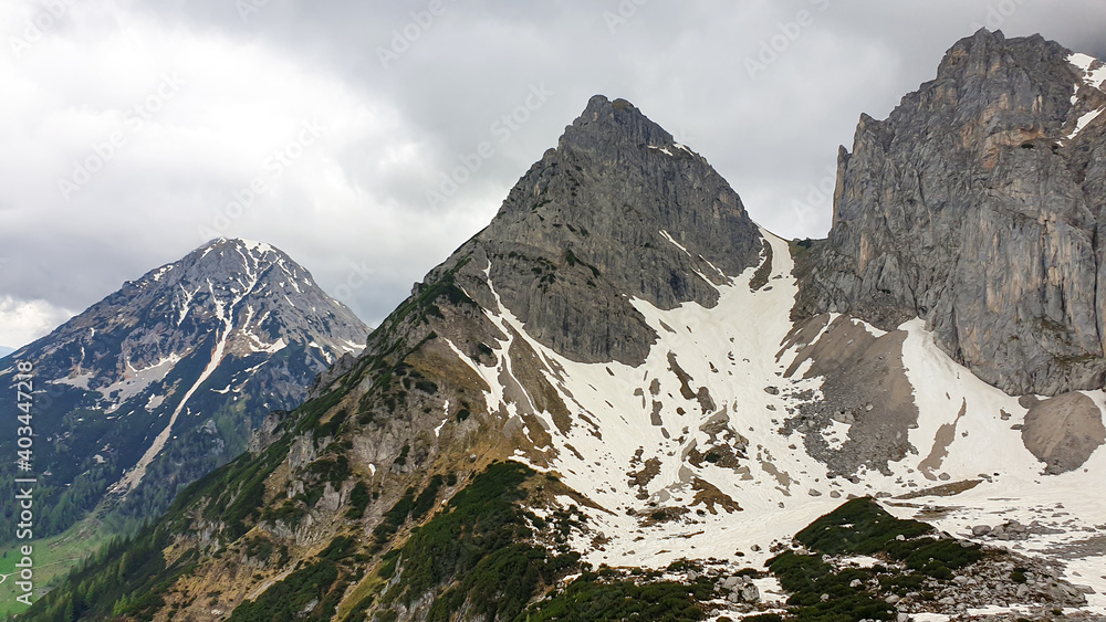 A panoramic view on the Alpine peaks in Austria from the Marstein. The slopes are still partially covered with snow. Stony and sharp mountains. Overcast. Baren slopes, green valley below. Serenity