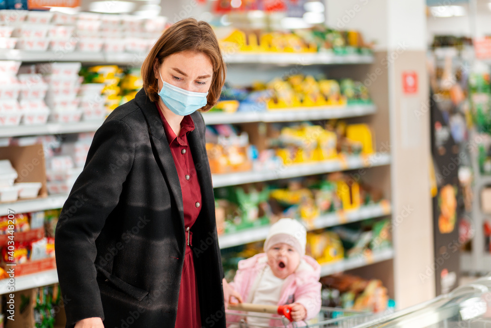 Shopping. A young mother in a medical mask chooses products. In the background, in a blur, there is a grocery cart with a baby sitting in it. The concept of a new normal