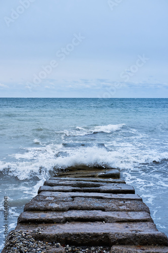 Waves break on rocks and fly apart into small drops and turn into foam. Long concrete breakwater stretching away into depths of stormy blue sea.