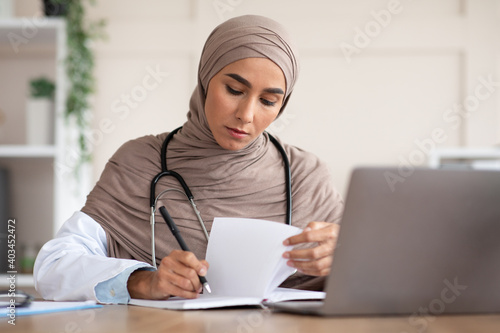Concentrated young woman doctor in hijab taking notes in clinic