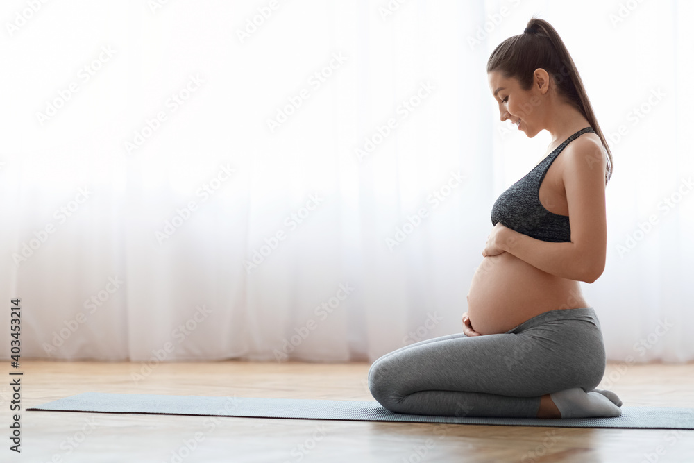 Pregnant woman training yoga at home, sitting on mat and touching belly
