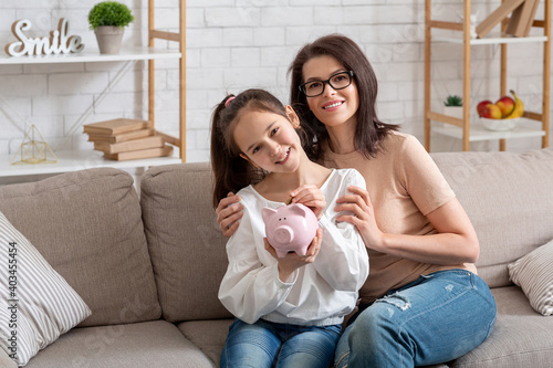 Fotografie Happy young mother and her teen daughter holding piggybank, sitting on sofa at h