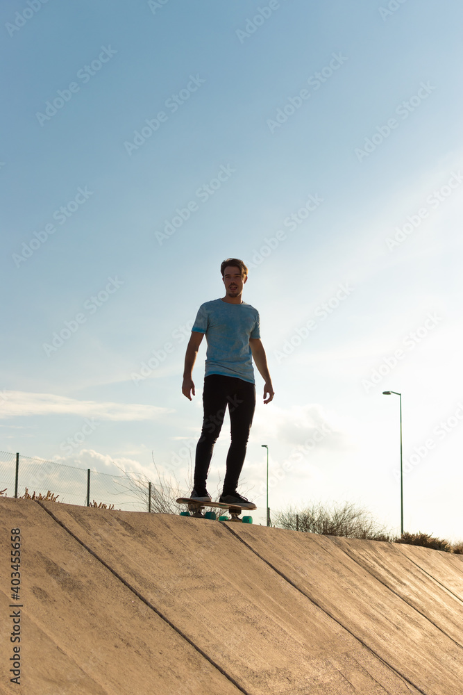 Young man wearing a blue t-shirt and black trousers riding a skateboard with a blue sky Young man wearing a blue t-shirt and black trousers riding a skateboard with a blue sky