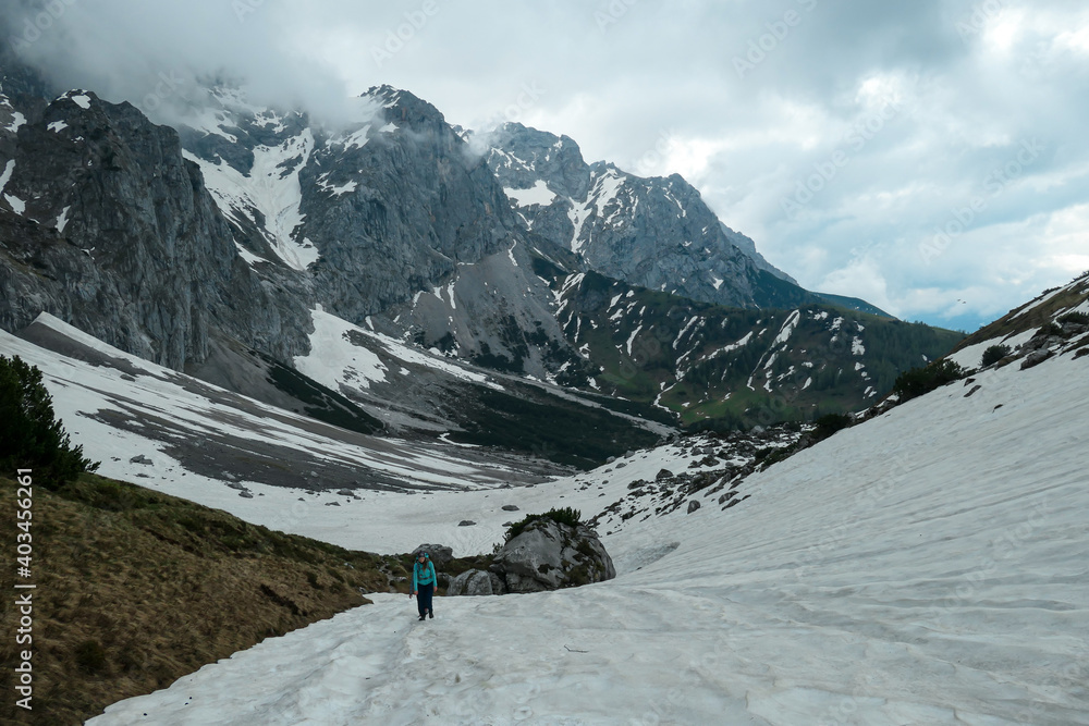A backpacker woman hiking through a snow covered slope of Marstein in Austrian Alps. Stony and sharp mountains. Overcast. Baren slopes, green valley below. Serenity and calmness. Hiking in the clouds