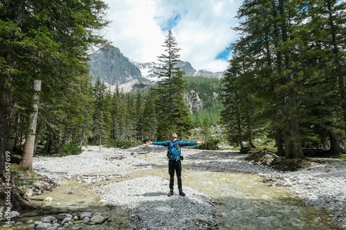 A backpacker man standing by a rushing torrent in region of Dachstein, Austria. There is dense forest behind, and high Alpine peaks. He is spreading his arms wide in gesture of freedom. Serenity
