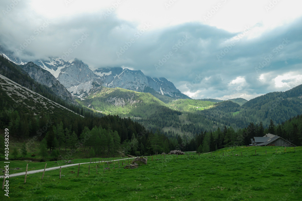 A gravelled road through Alpine valley in Austria in the region of Dachstein. High mountain around, partially covered with snow. Stony and sharp mountains. Overcast. Dense forest at the foothill.