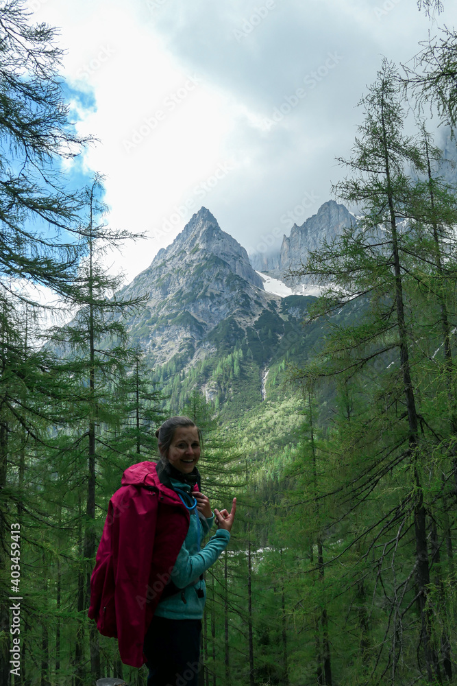 Naklejka premium A backpacker woman in pink jacket pointing to the peak of Marstein, Austrian Alps. The slopes are partially covered with snow. Stony and sharp mountains. Overcast. Dense forest at the foothill. Happy