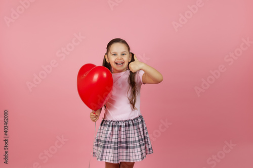 close-up glad, joyful child in a pink dress holds a red balloon in the shape of a heart and gives a thumbs up, isolated on a pink background. © Юля Бурмистрова